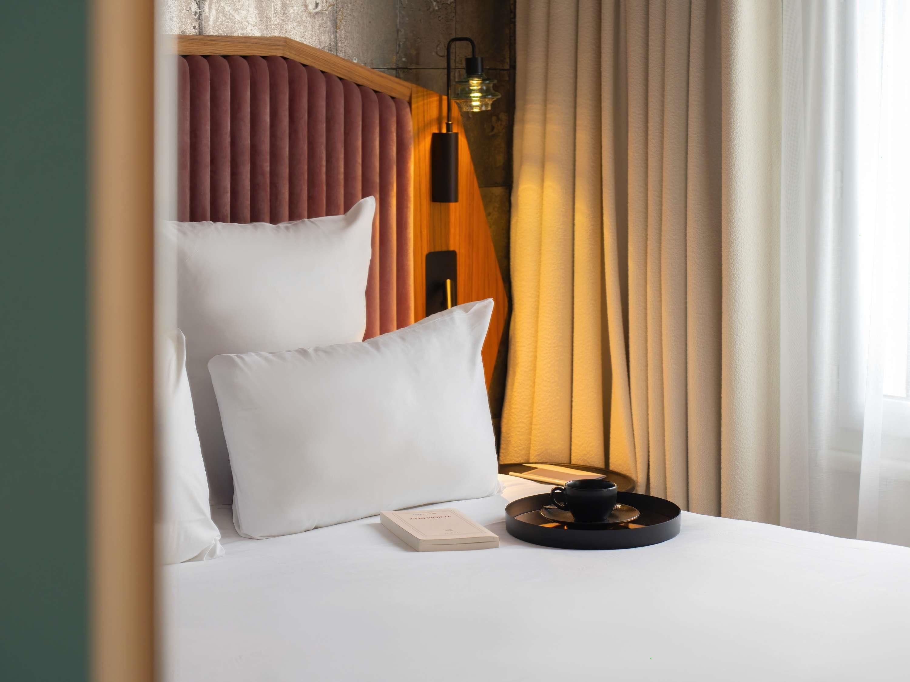 Een hotelkamer met een bed, kussens, een nachtkastje met een kopje en gordijnen.
