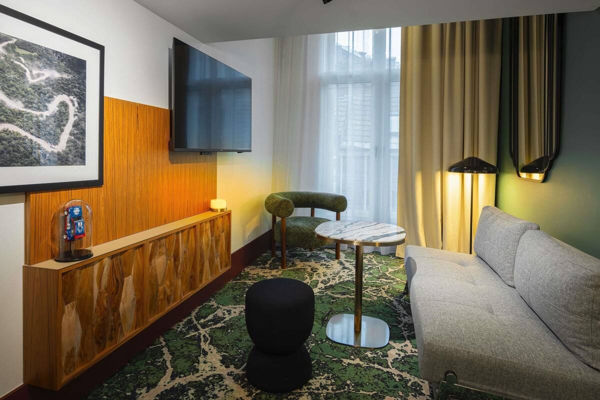 Una habitación de hotel moderna con un mostrador de recepción de madera, un televisor de pantalla plana, una alfombra estampada verde, una mesa redonda, una silla verde, un sofá gris y un ventanal con cortinas.