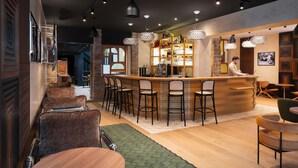 A bar area with a wooden counter, bar stools, and a bartender. There are hanging pendant lights and a bar shelf with bottles. - Hotel Maison Louise Lille Centre - Handwritten Collection (Lille)