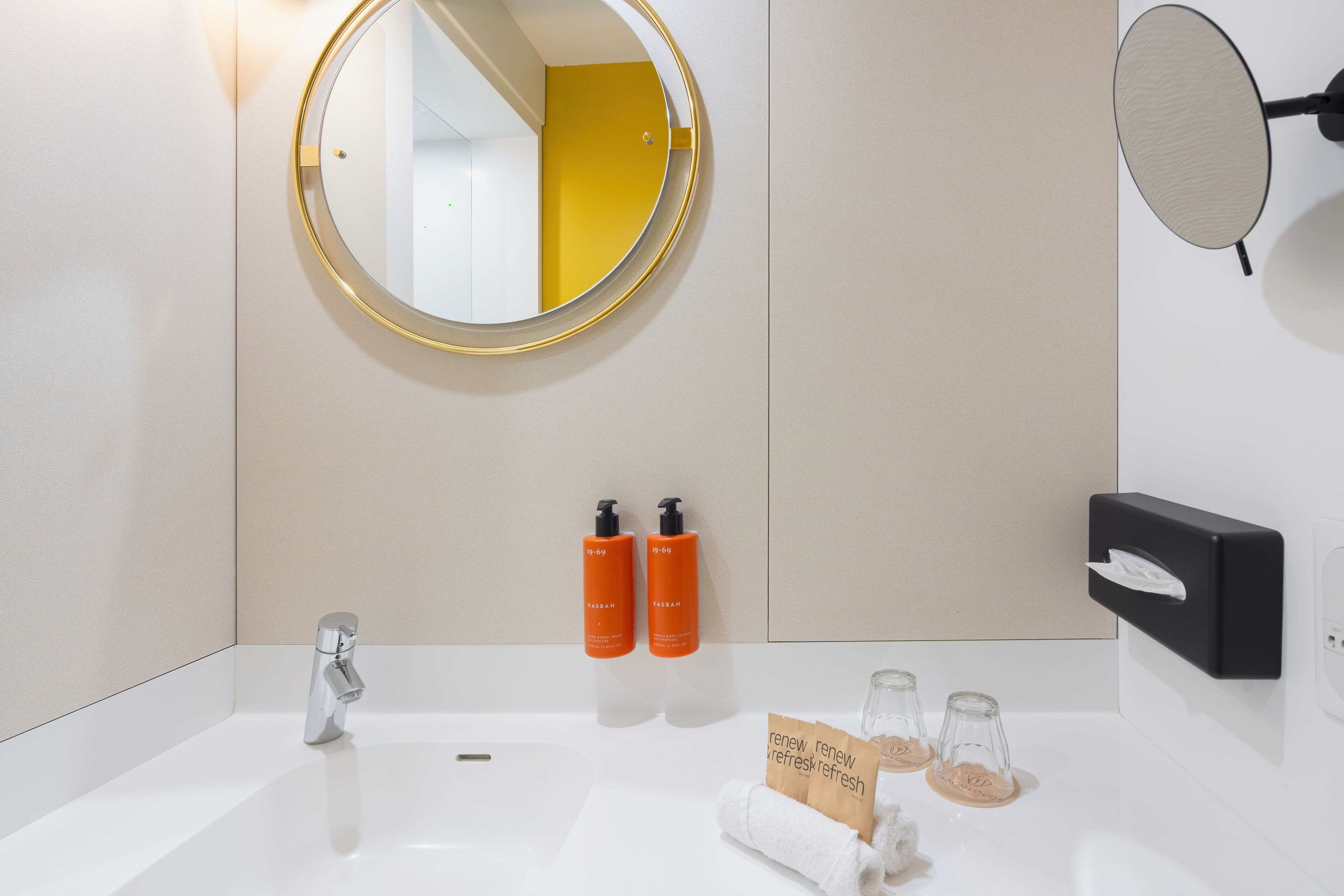 A bathroom with a round mirror, a handheld mirror, a sink, two orange Kasbah body wash bottles, a black soap dispenser, and two glass containers with brown labels.