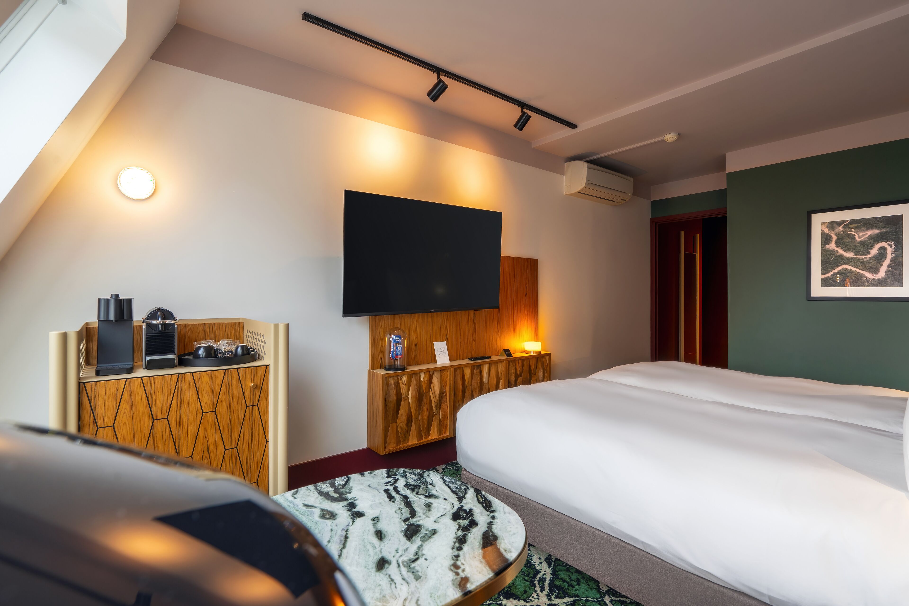 A modern hotel room with a large bed, a flat-screen TV mounted on the wall, a wooden side table with a coffee maker, and a small round table with a patterned tablecloth.