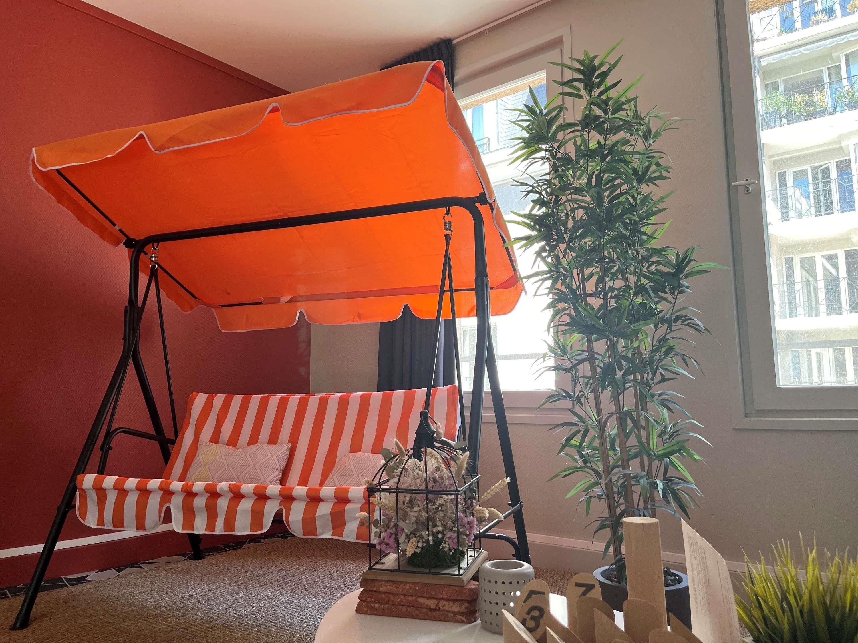 Une pièce avec un mur rouge, un fauteuil suspendu doté d’un auvent orange, une plante en pot et une fenêtre donnant sur des immeubles.