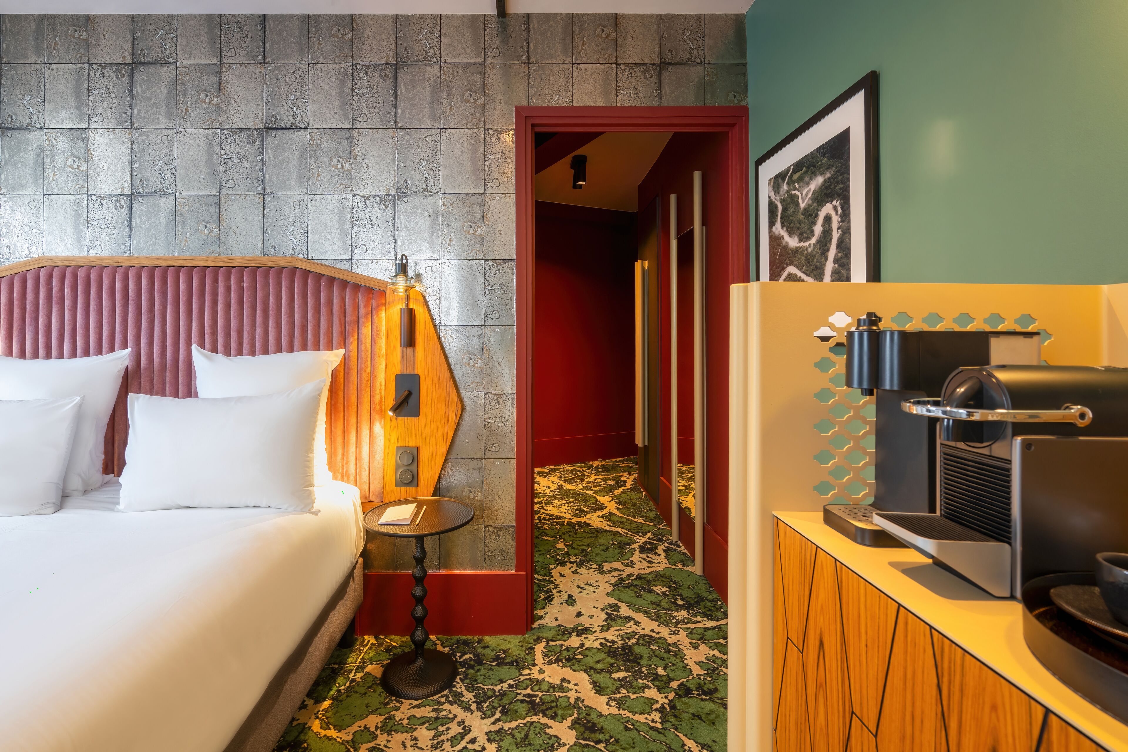 A hotel room with a bed, a small table with a lamp, a coffee machine, and a red door leading to a hallway.