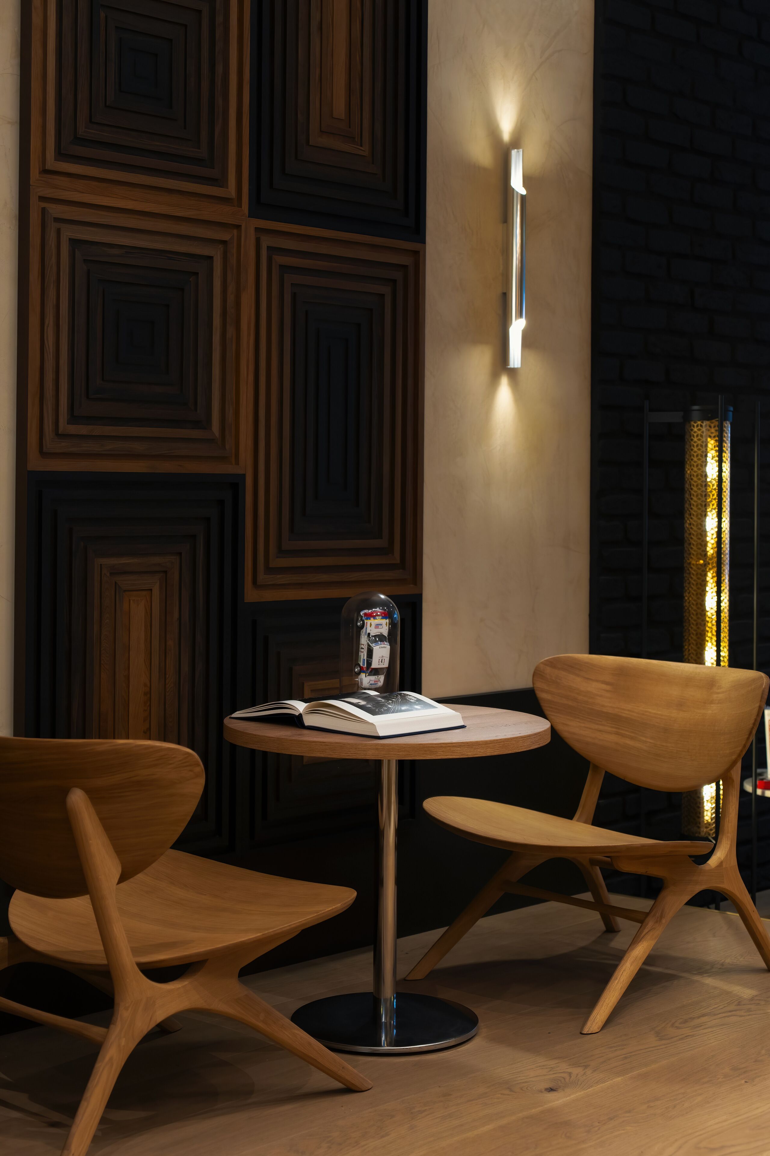 A modern interior with a round table and two wooden chairs, a wall with geometric patterns, and a tall, narrow light fixture.