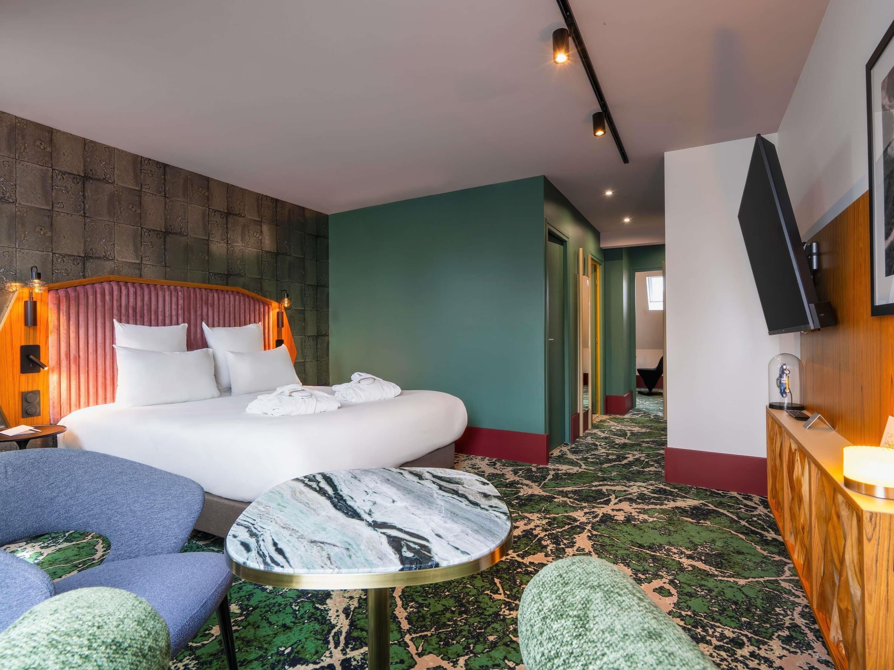 A hotel room with a bed, a round table, a chair, a TV, and a hallway.