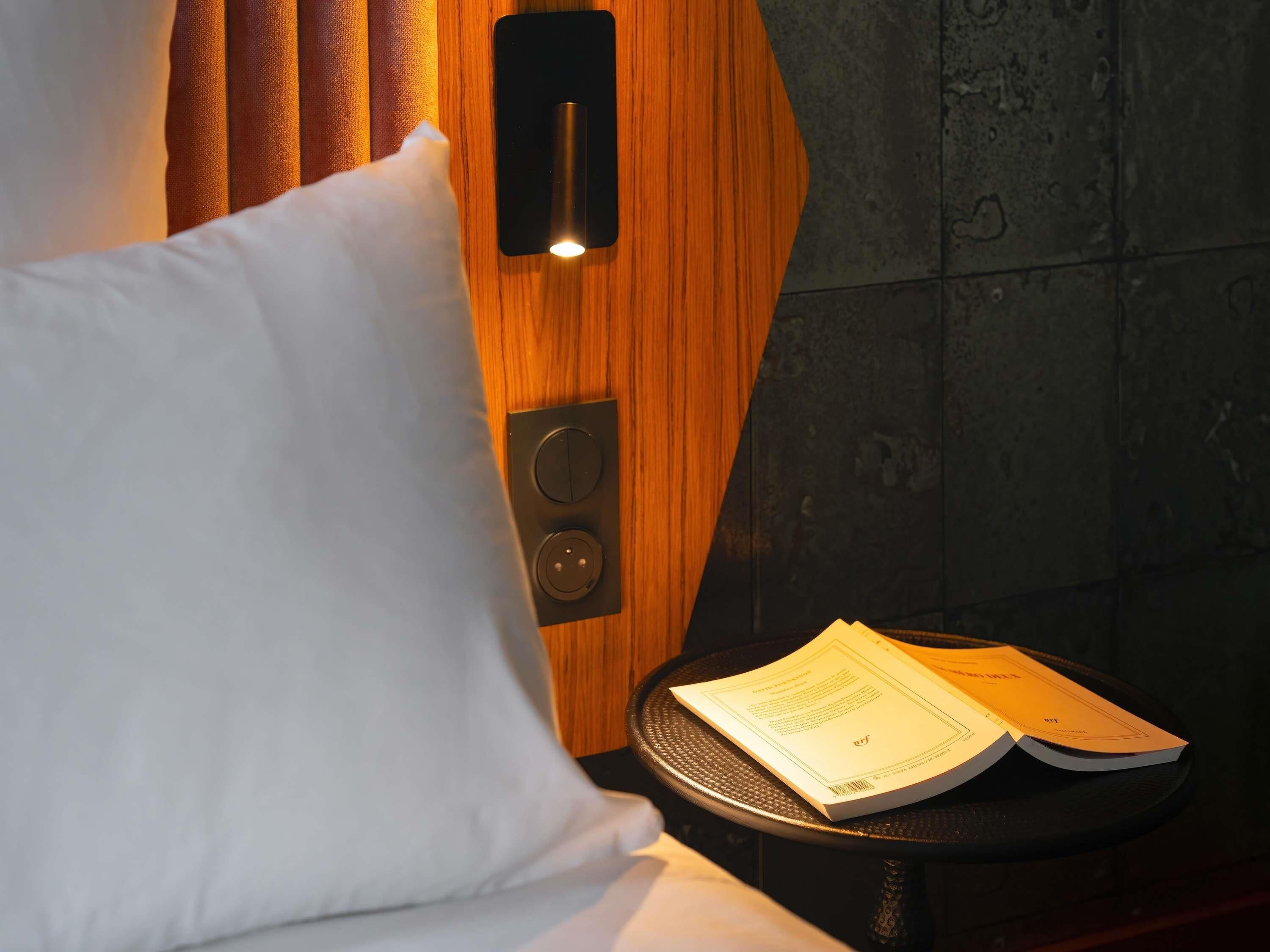 A hotel room with a wooden panel wall, a bed with white bedding, a bedside table with a book, and a wall-mounted light switch.