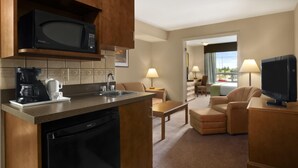 Habitación de hotel con kitchenette, que incluye microondas, cafetera y lavavajillas. Cuenta con una zona de estar con sofá y sillones, y un televisor sobre un mueble.