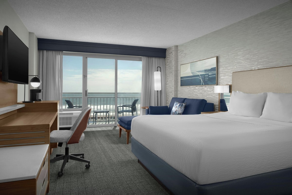 A hotel room with a large bed, a desk with a chair, a TV, and a balcony with a view of the ocean.
