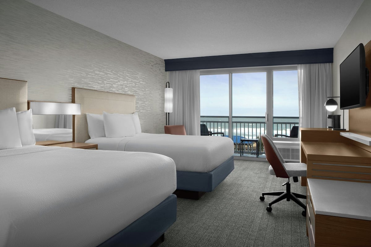 A hotel room with two beds, a desk, a chair, a TV, and a balcony with a view.
