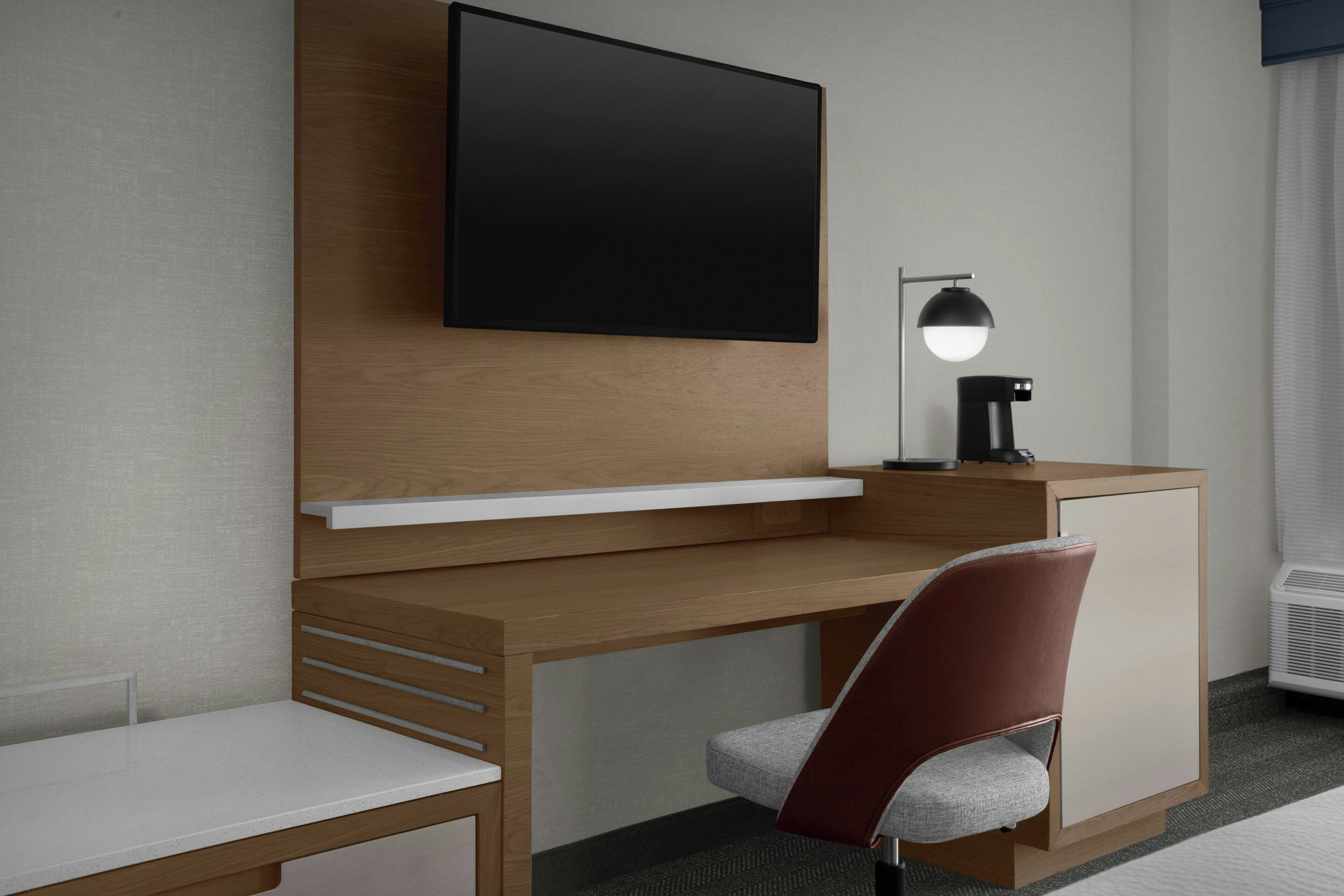 A modern hotel room with a wooden desk, a flat-screen TV, a desk lamp, and a cushioned chair.