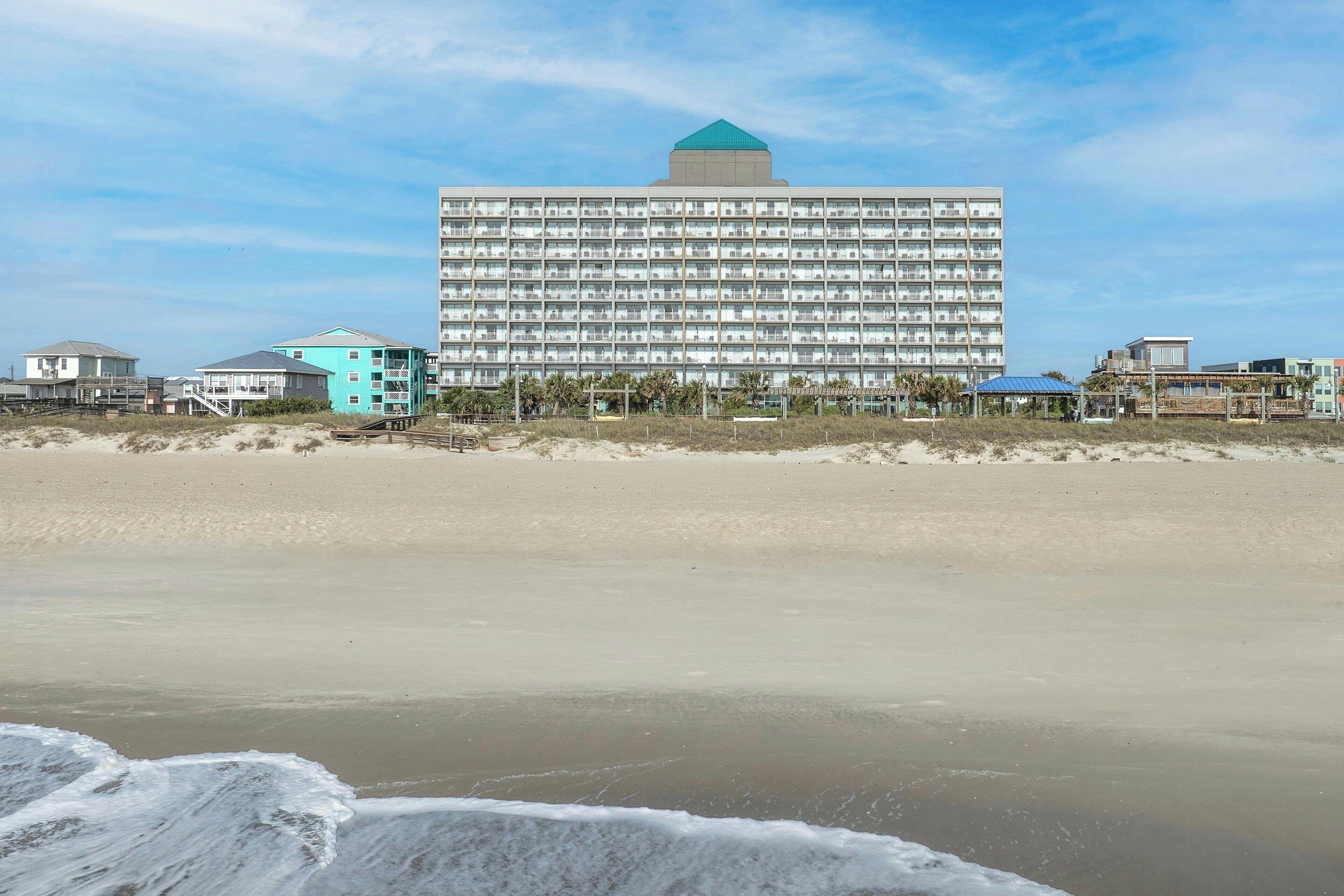 A large hotel with a flat roof and multiple balconies, situated on a beach with sandy dunes and a clear sky.