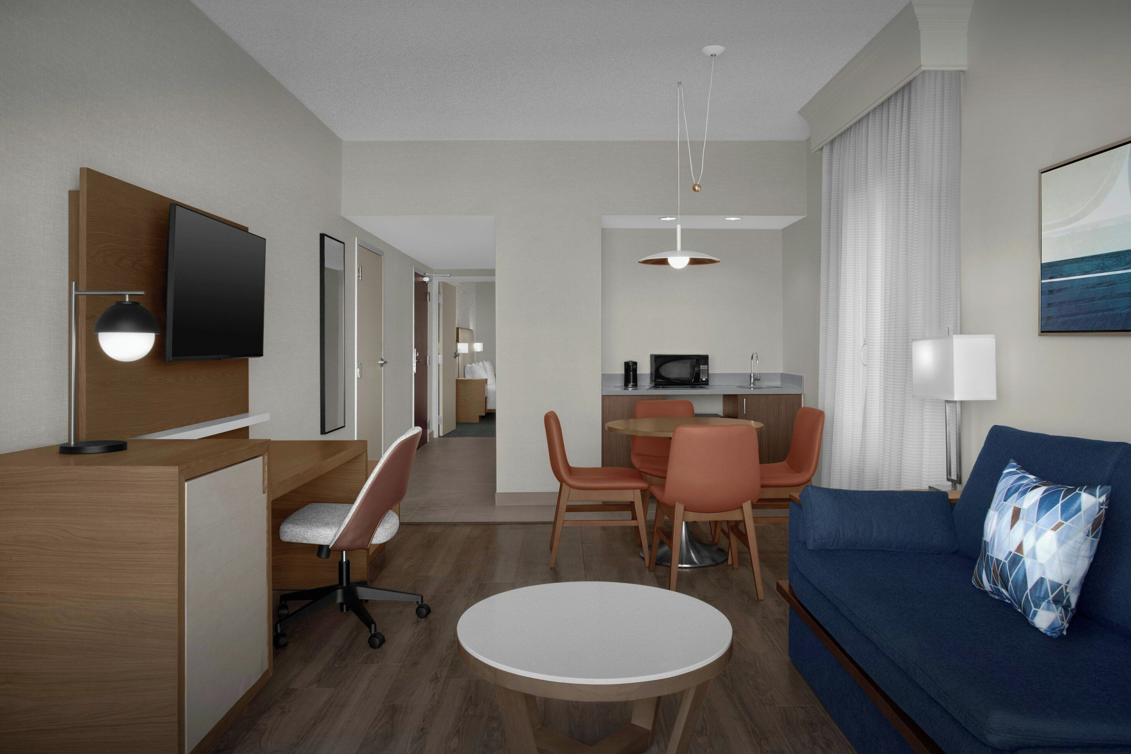 Quarto de hotel moderno com escrivaninha de madeira, mesa de jantar redonda, sofá azul e televisão fixada na parede.