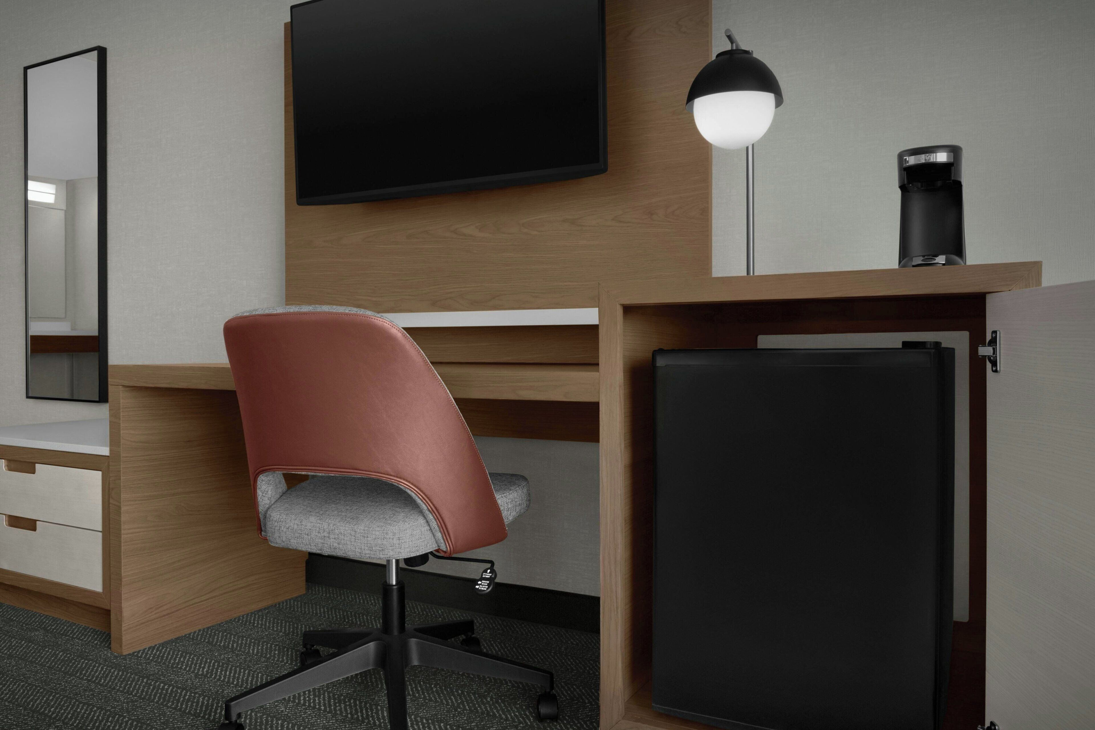 A modern hotel room with a wooden desk, a red office chair, a flat-screen TV, a desk lamp, and a coffee maker.