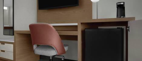 A modern hotel room with a wooden desk, a red office chair, a flat-screen TV, a desk lamp, and a coffee maker.