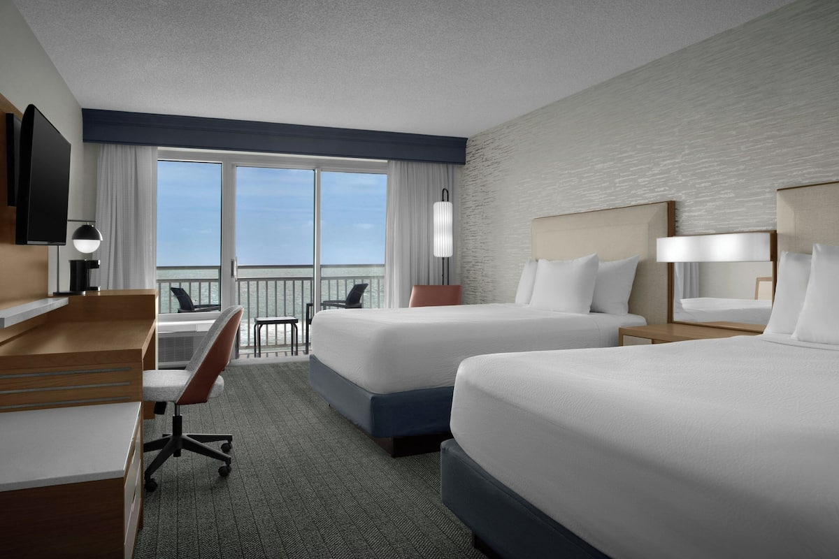 A hotel room with two beds, a desk, a chair, a TV, and a balcony with a view.