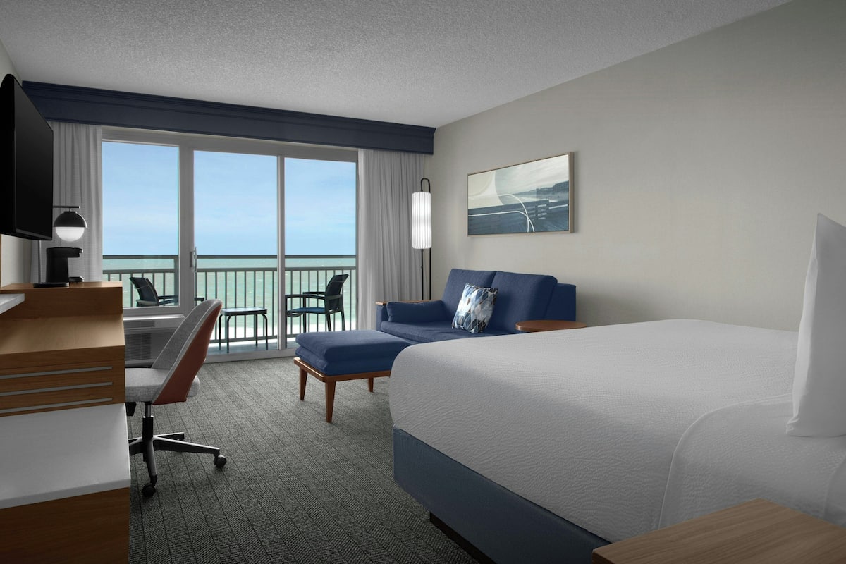 A hotel room with a bed, desk, chair, and a view of the ocean.