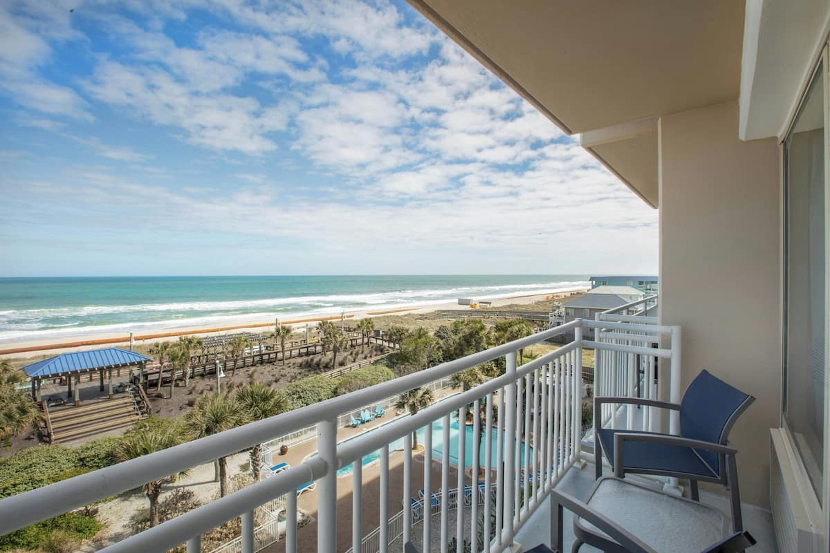 A balcony with a view of the beach and ocean, a pool, and outdoor seating.