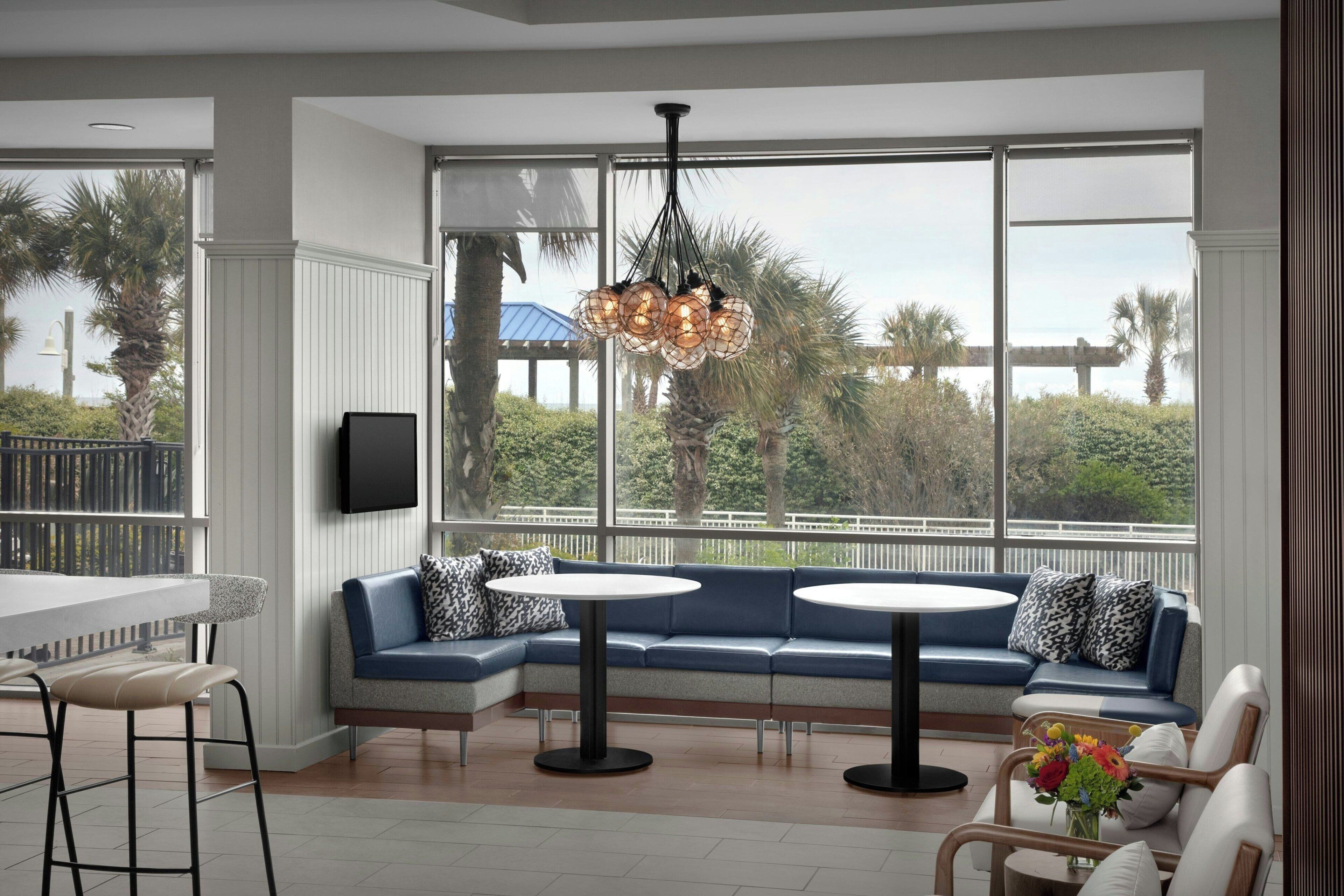 A modern interior with a sectional sofa, round tables, and a large window with a view of palm trees and a bridge.