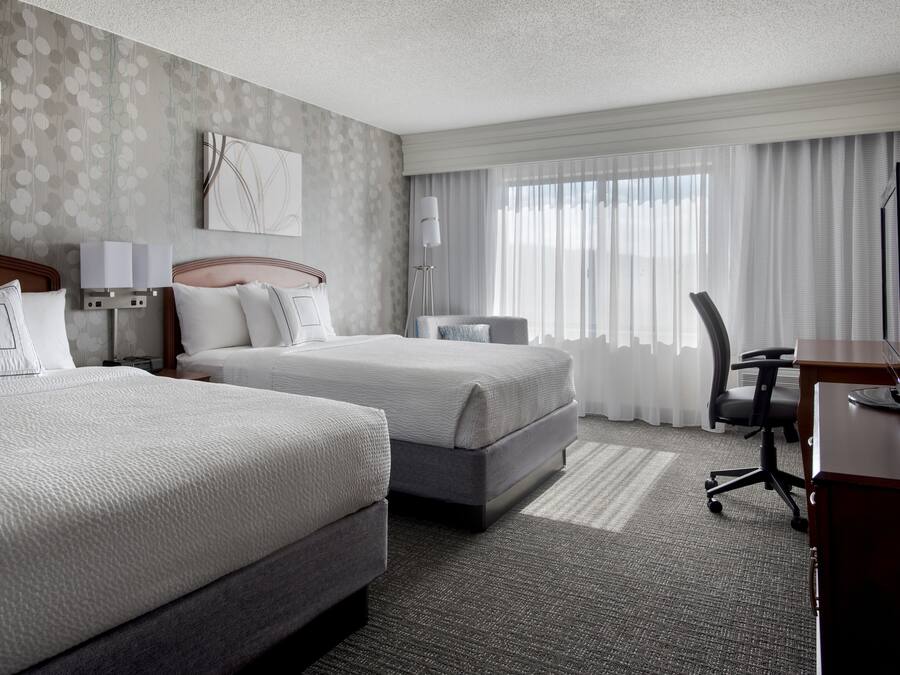 A hotel room with two beds, a desk, a chair, and a lamp.