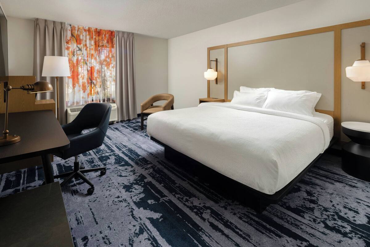Una habitación de hotel con una cama grande, un escritorio con lámpara, una silla, un espejo amplio y una ventana con cortinas.