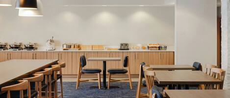 A modern dining area with wooden tables and chairs, a kitchen in the background, and pendant lighting.