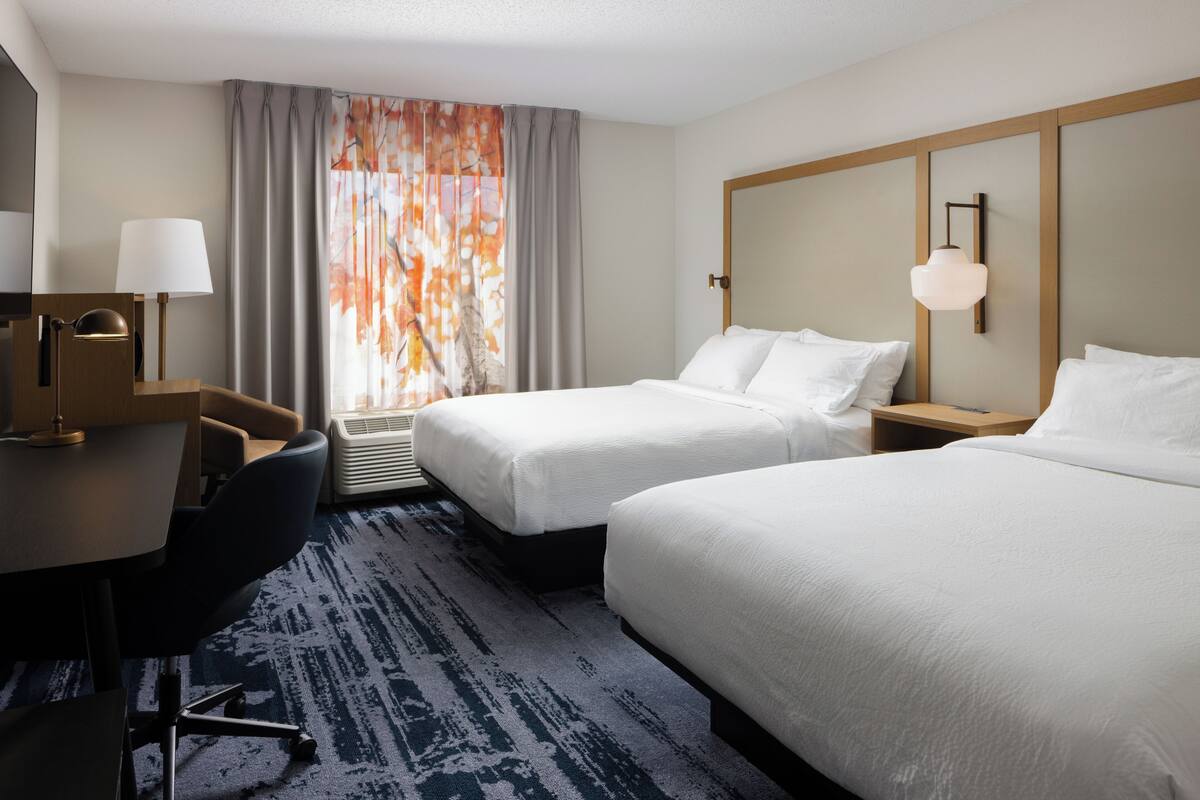 Habitación de hotel con dos camas, un escritorio con silla, una lámpara, luz en la pared y una ventana con cortinas.