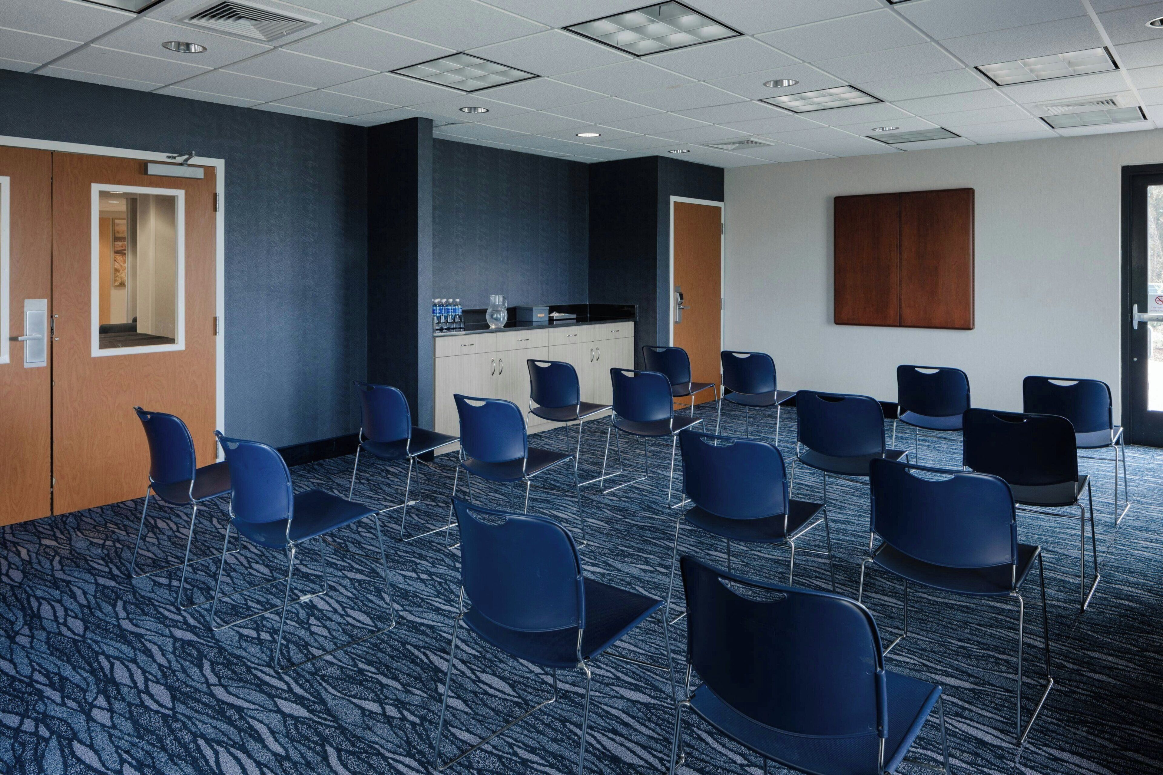 A conference room with a carpeted floor, rows of chairs, a kitchenette, and a door.