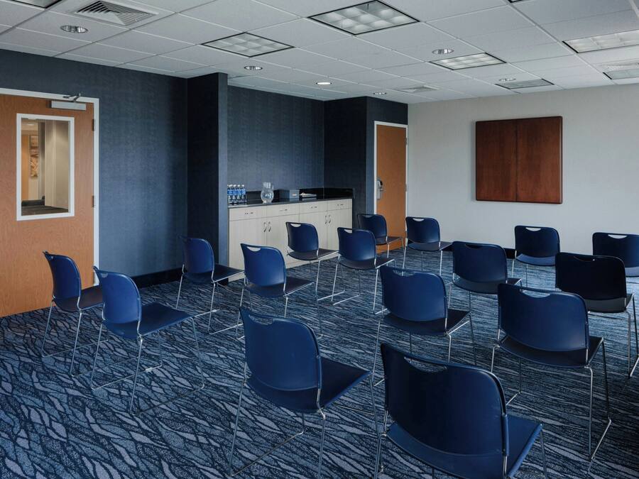 A conference room with a carpeted floor, rows of chairs, a kitchenette, and a door.