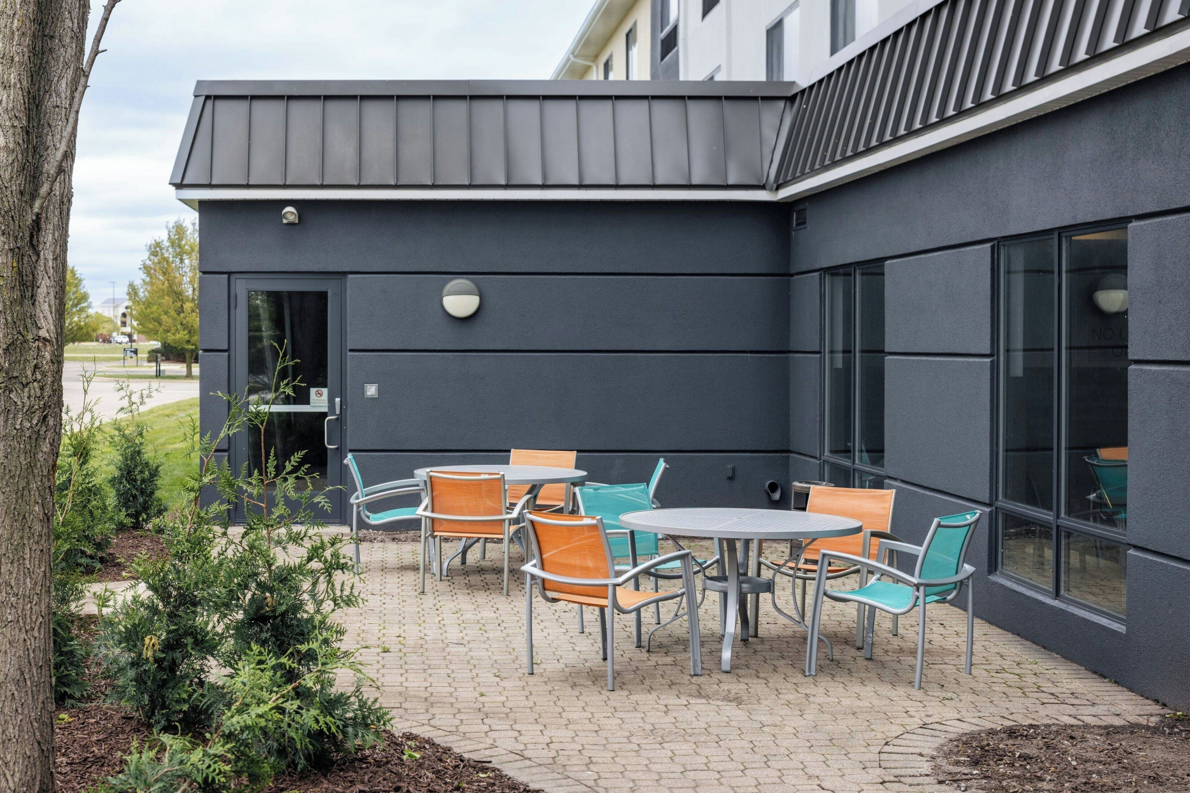 An outdoor patio with metal tables and chairs, a small garden area, and a building with large windows and a dark facade.