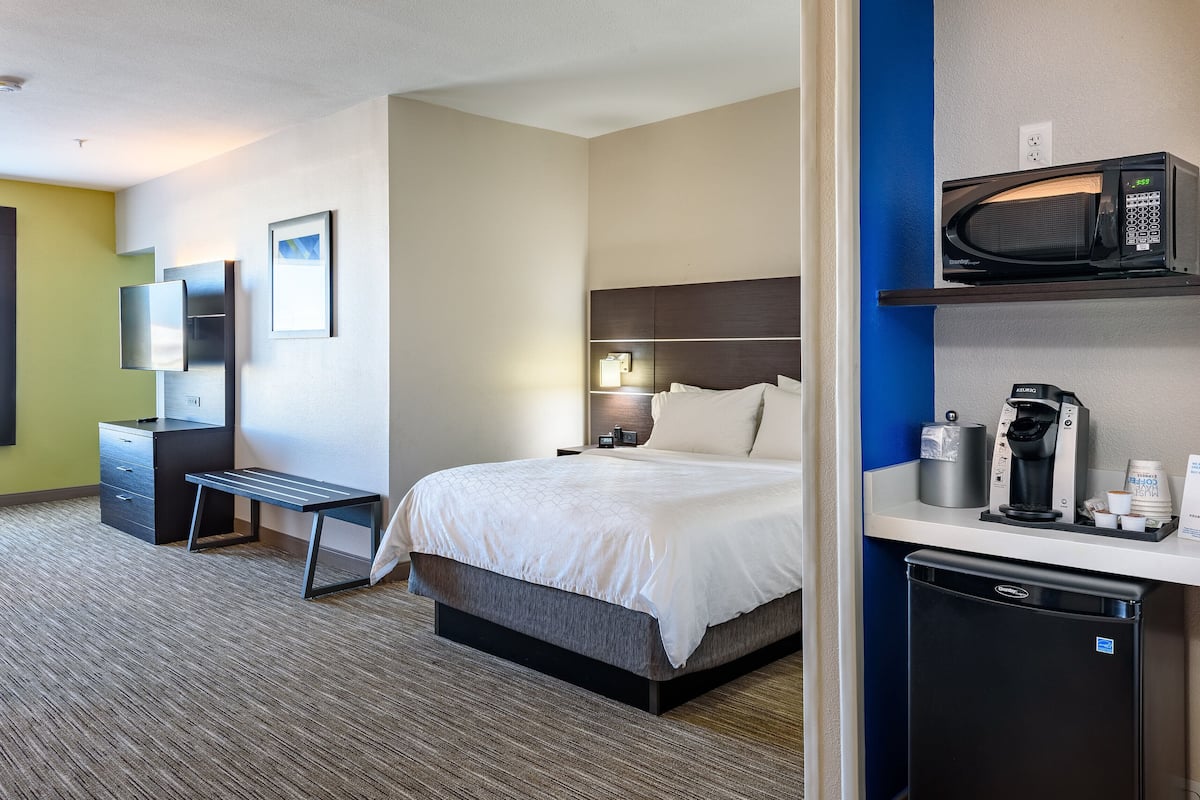 Quarto de hotel com cama, mesa de cabeceira, escrivaninha, cadeira, televisão e máquina de café.