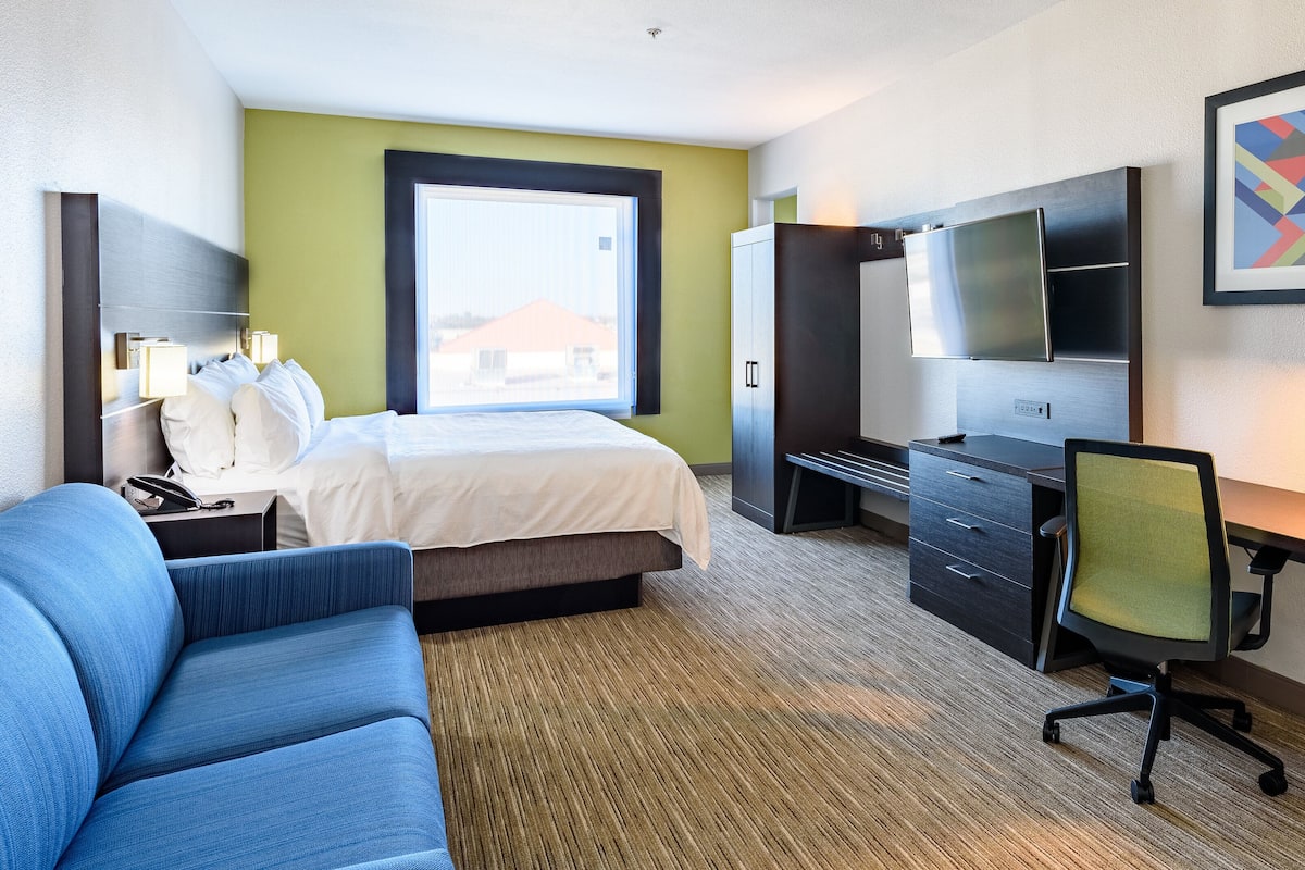 Quarto de hotel com cama, escrivaninha, cadeira, televisão e quadro na parede.