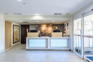 A hotel reception desk with a digital check-in kiosk, a framed artwork on the wall, and a glass door entrance.