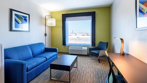 A modern hotel room with a blue sofa, a small table, a chair, a large window with a view, and a wall-mounted air conditioner. - Holiday Inn Express Hotel & Suites Bastrop by IHG (Bastrop)