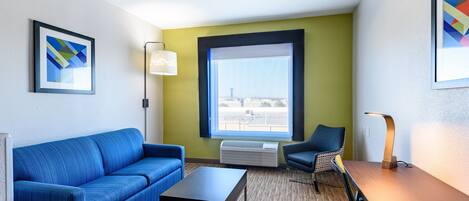 A modern hotel room with a blue sofa, a small table, a chair, a large window with a view, and a wall-mounted air conditioner.