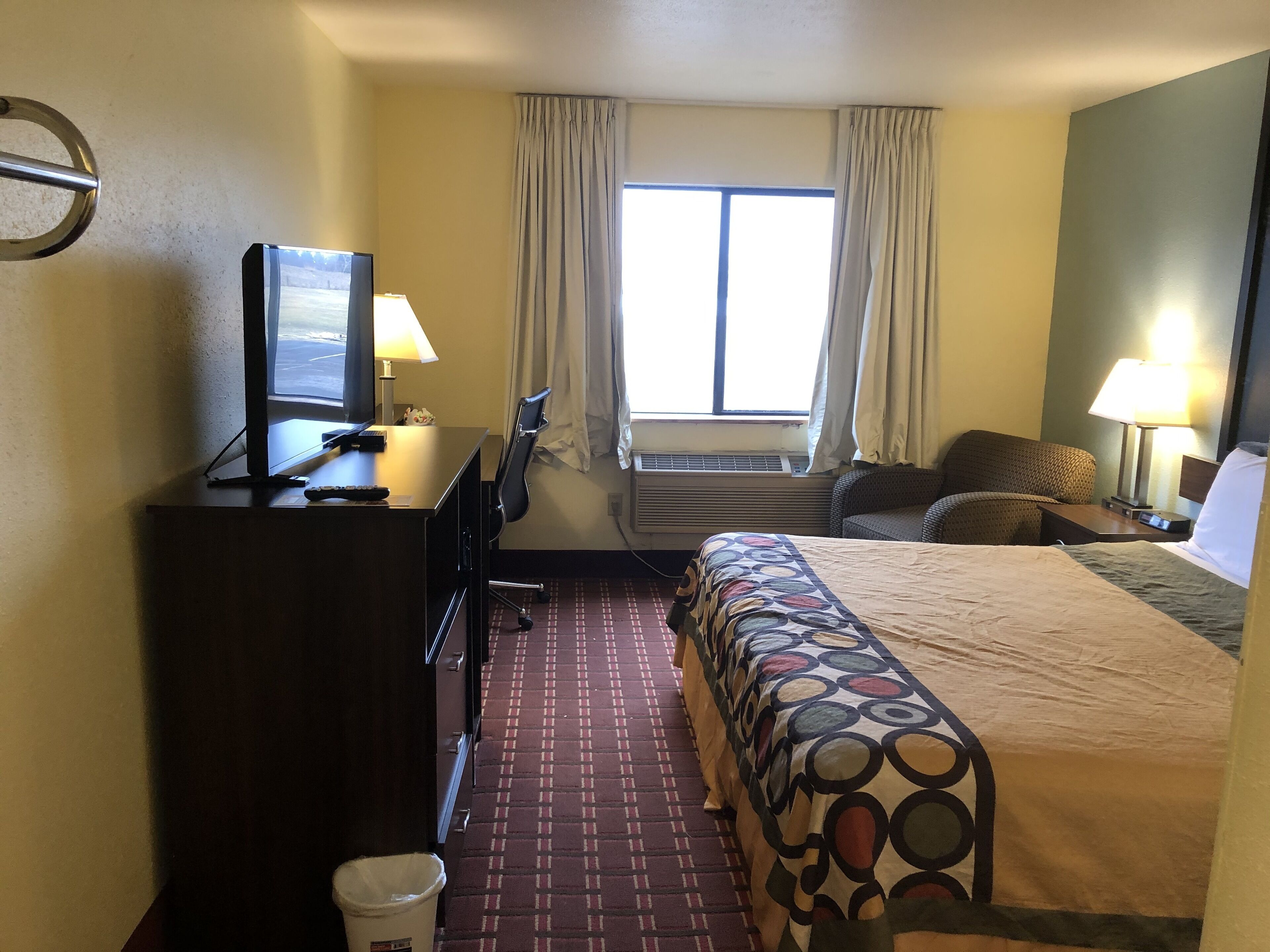 A hotel room with a bed, a desk with a computer and lamp, a chair, and a window with curtains.