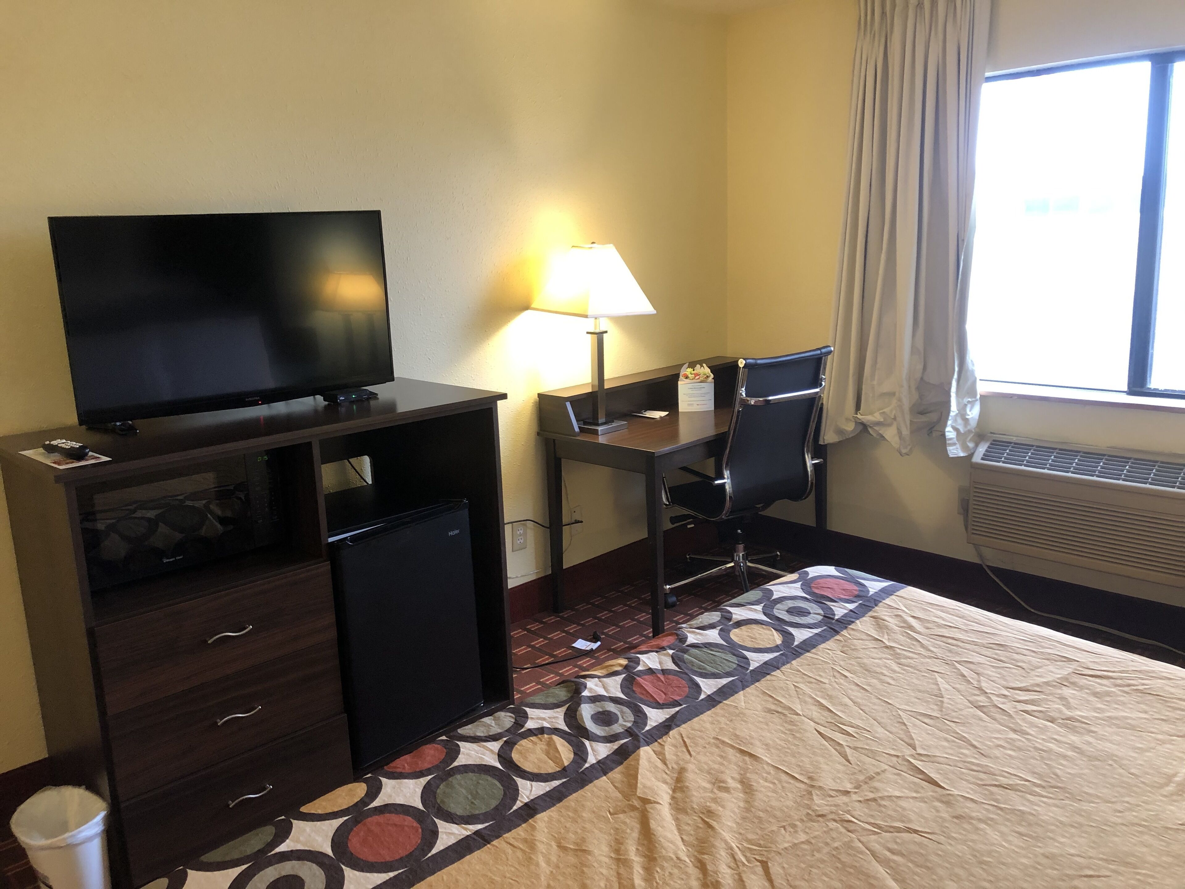 A hotel room with a TV, desk, chair, lamp, air conditioner, and a patterned rug.