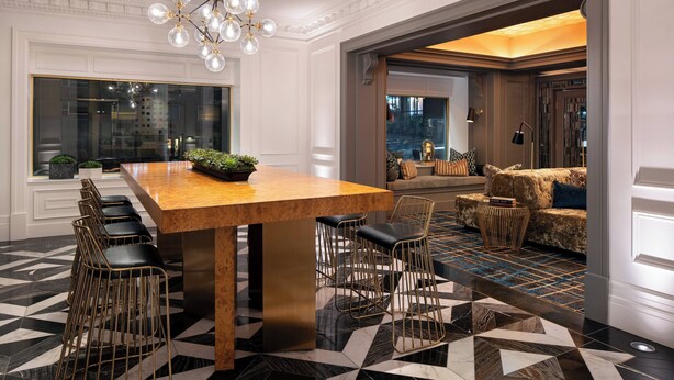 Een moderne hotellobby met een lange houten bar, barstoelen, een vloer met een patroon en een zithoek met een bank en kussens.