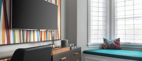 A modern bedroom with a large flat-screen TV, a desk with drawers, a window seat with a colorful cushion, and a striped wall art piece.