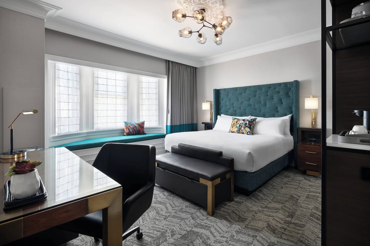 Une chambre d’hôtel moderne avec un grand lit, un bureau avec une chaise, un banc, une lampe de bureau et une fenêtre avec des rideaux.