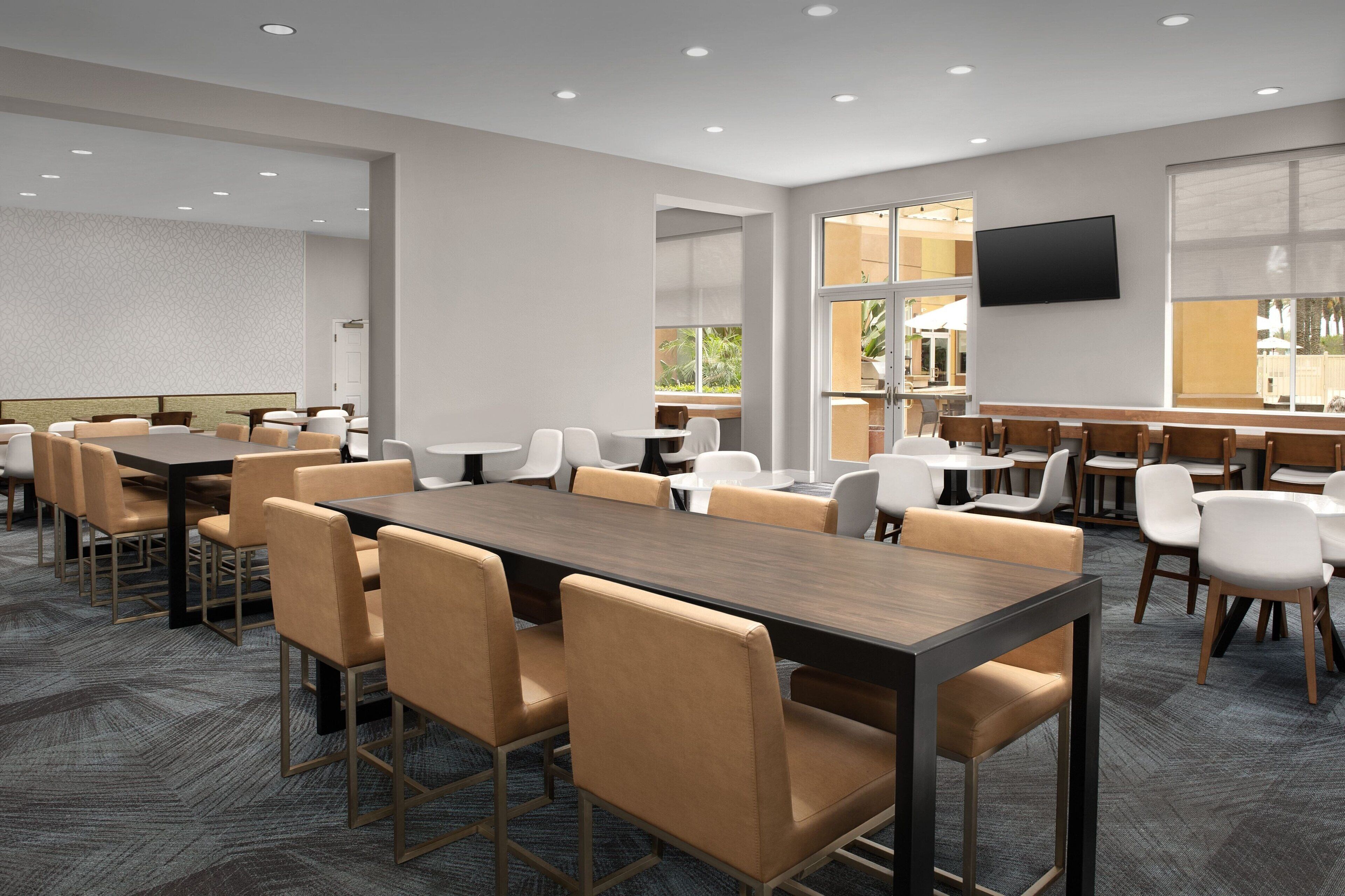 A modern hotel dining area with multiple tables and chairs, a flat-screen TV mounted on the wall, and large windows providing natural light.