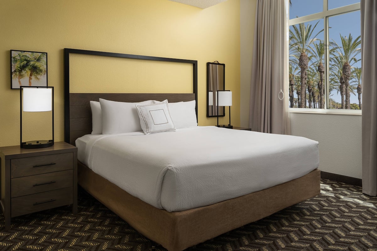 Une chambre d’hôtel avec un grand lit, deux tables de chevet, une commode, une fenêtre donnant sur des palmiers et un tableau au mur.