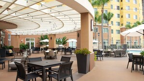 An outdoor patio area with tables and chairs, surrounded by a pool and a building with multiple windows. - Residence Inn By Marriott Anaheim Resort Area (Garden Grove)