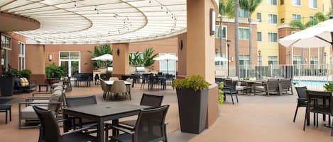 Un espace extérieur aménagé en terrasse, avec des tables et des chaises, entouré d’une piscine et d’un bâtiment doté de nombreuses fenêtres.