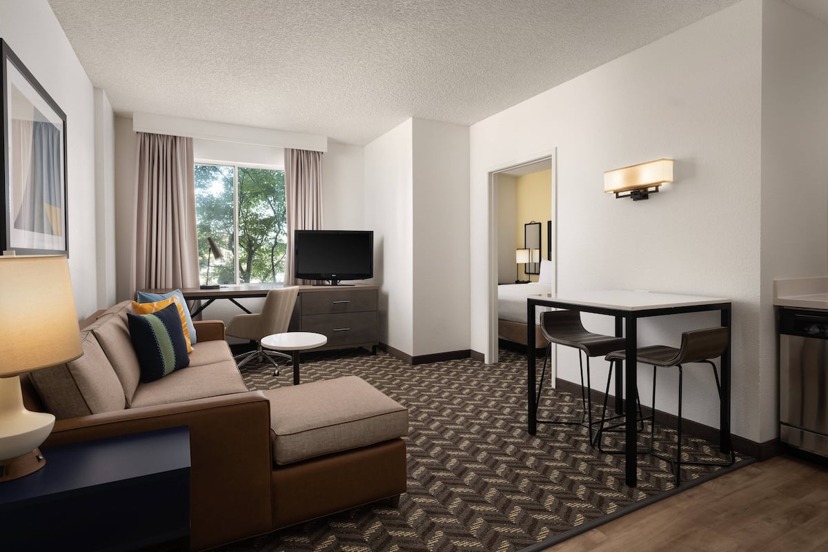 Une chambre d’hôtel comprenant un canapé, un téléviseur, une table à manger avec des chaises, un petit coin cuisine et une grande fenêtre avec des rideaux.