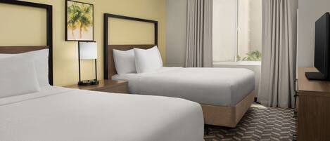 A hotel room with two beds, a nightstand with a lamp, a television, and a window with curtains.