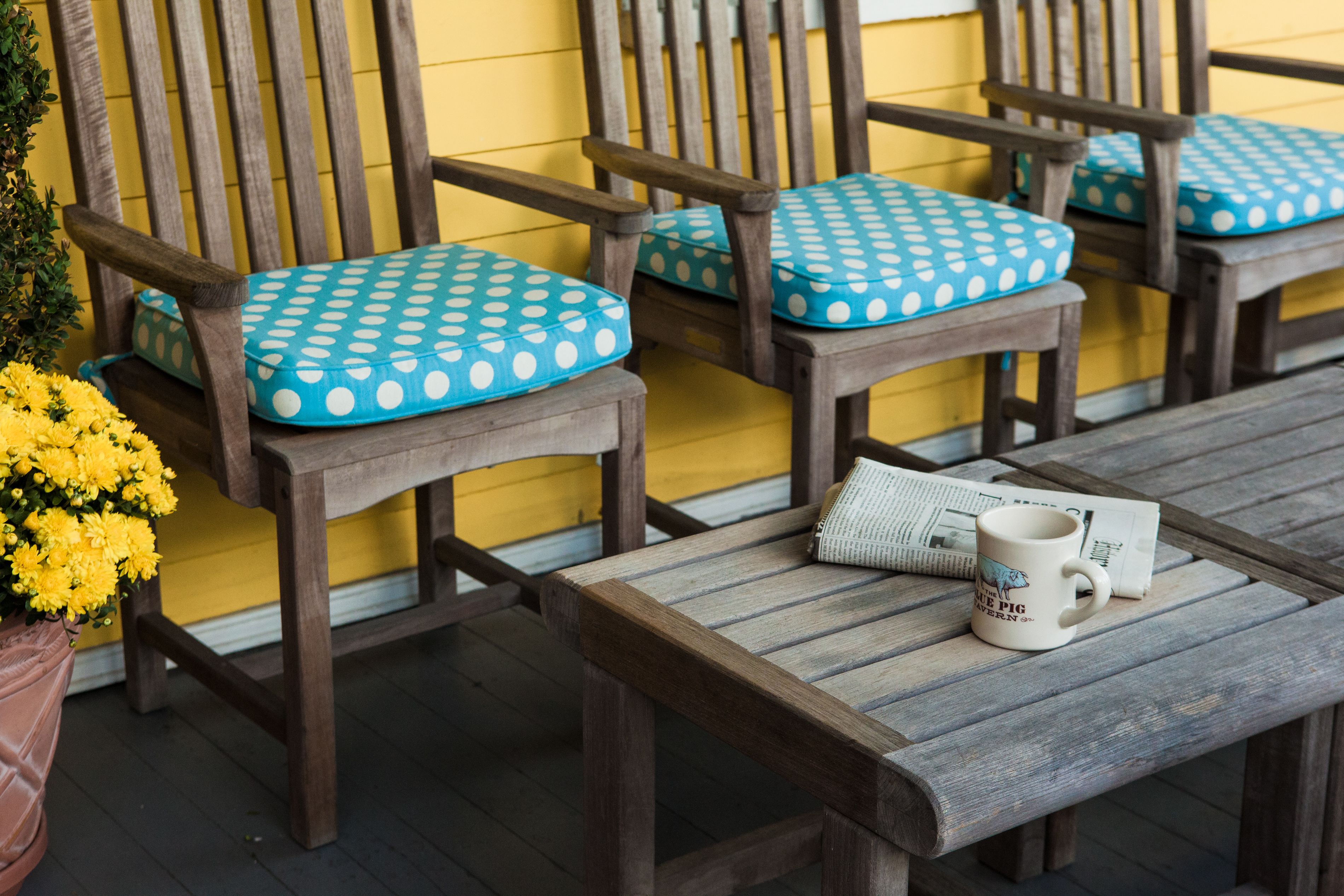 A wooden bench with a polka dot cushion, a wooden table with a mug and newspaper, and a yellow wall in the background.