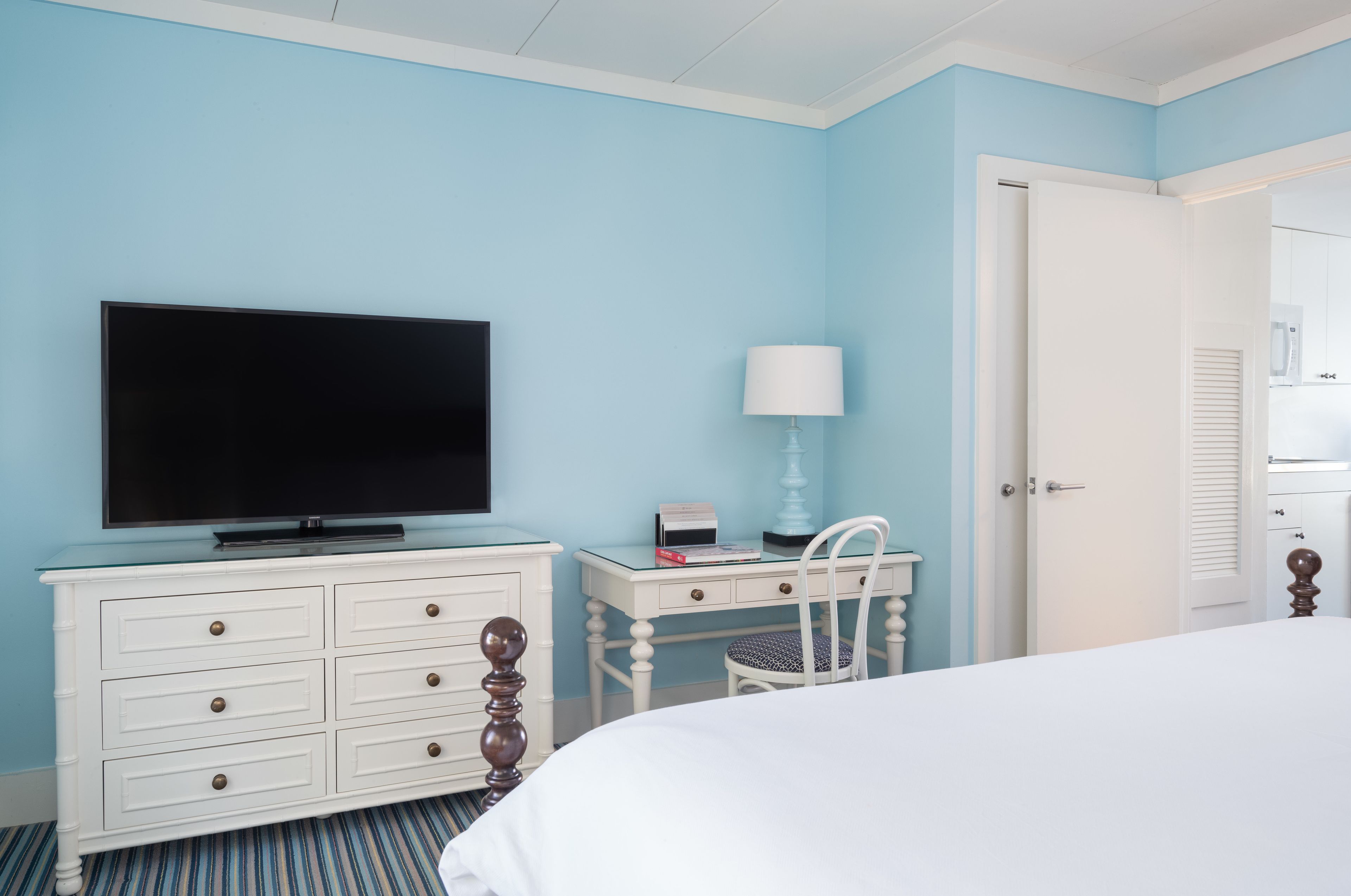 A bedroom with a bed, a TV on a dresser, a desk with a chair, and a door.