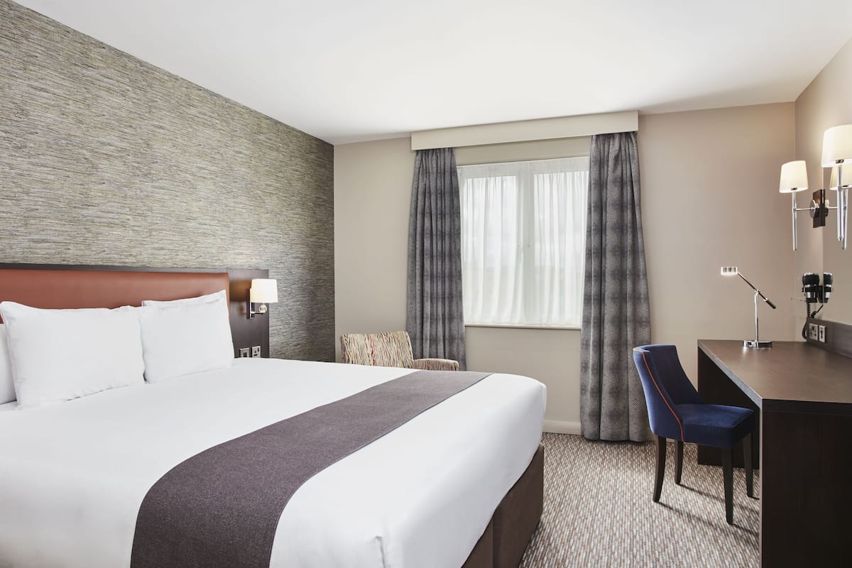 A hotel room with a large bed, a desk with a chair, a lamp, and a window with curtains.