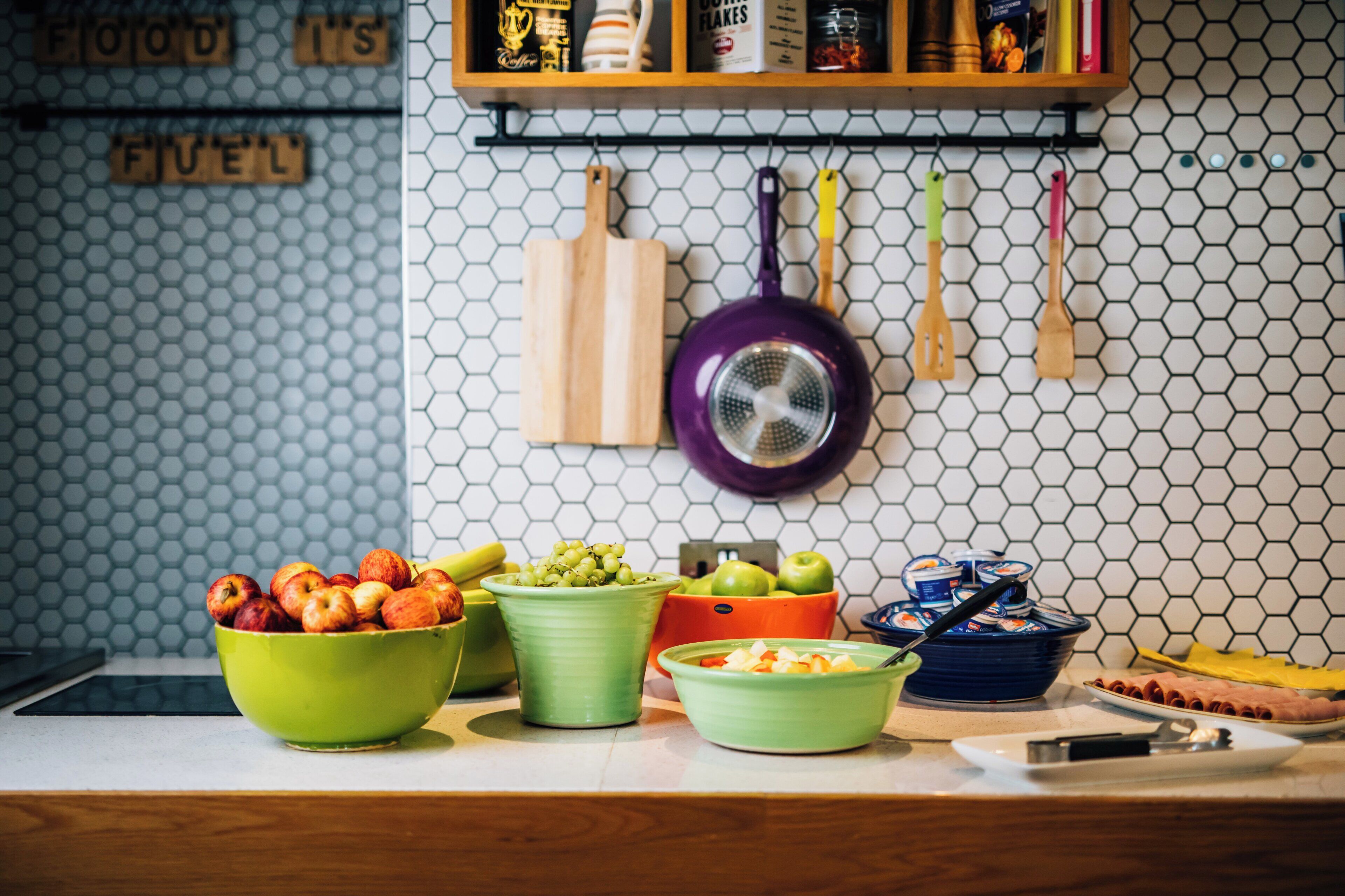 Una encimera de cocina con varios recipientes de frutas y verduras, una olla morada y una tabla de cortar de madera.