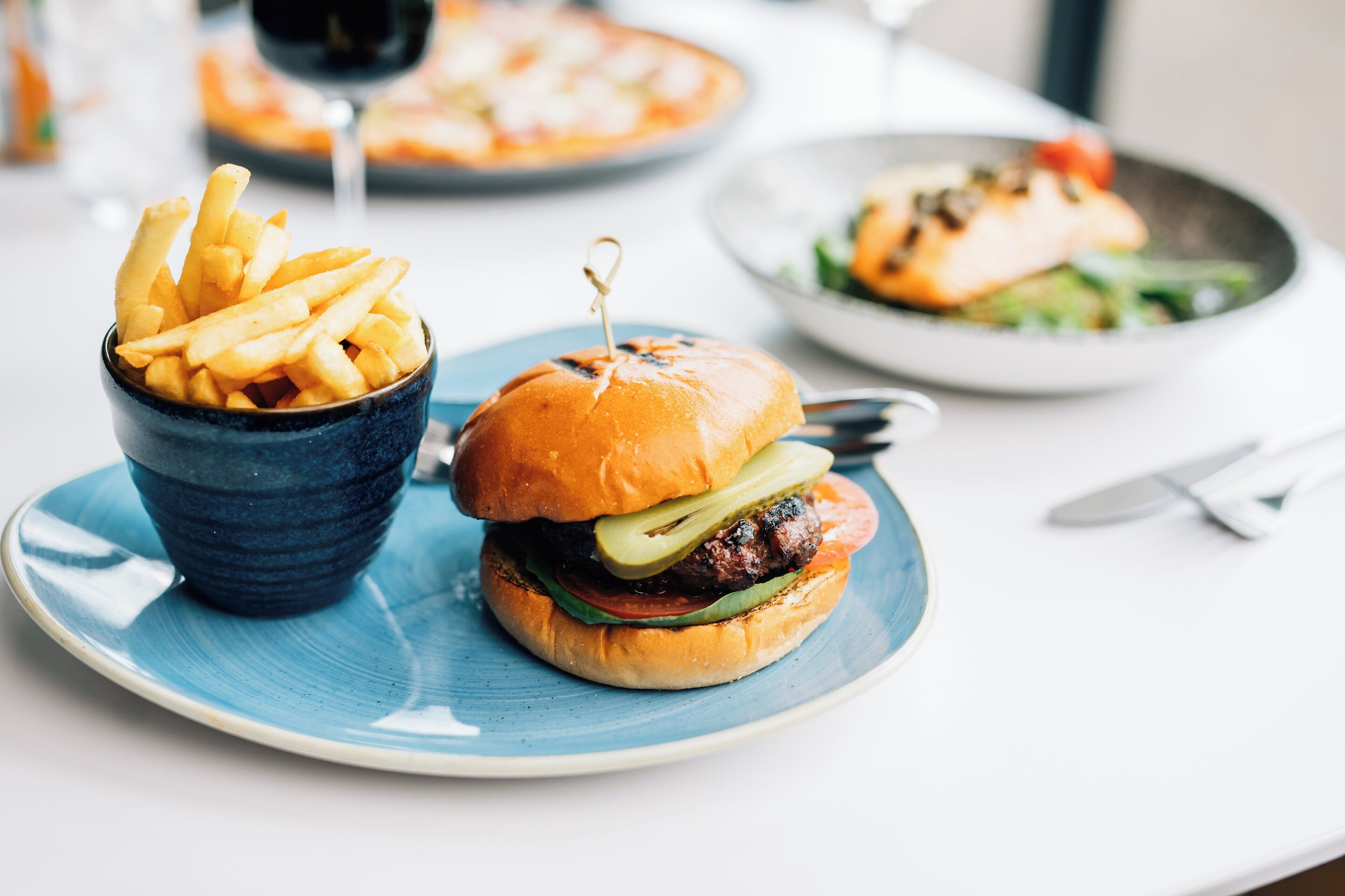 En tallerken med en burger og pommes frites, en skål med pommes frites og et glas vin på et bord.